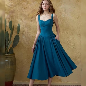 May include: A teal-colored dress with a sweetheart neckline and wide shoulder straps. The dress features a fitted bodice, a flared skirt, and pockets. The model is wearing gold-colored heels.