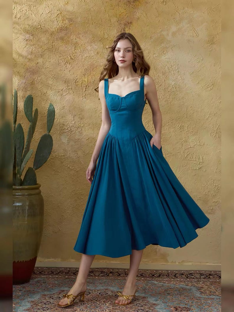 May include: A teal-colored dress with a sweetheart neckline and wide shoulder straps. The dress features a fitted bodice, a flared skirt, and pockets. The model is wearing gold-colored heels.