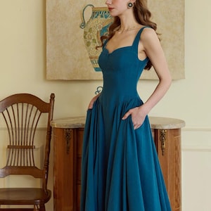 May include: A teal-colored dress with a sweetheart neckline and wide straps. The dress has a fitted bodice and a flared skirt with pockets. The model is standing in front of a painting of a vase and fruit.