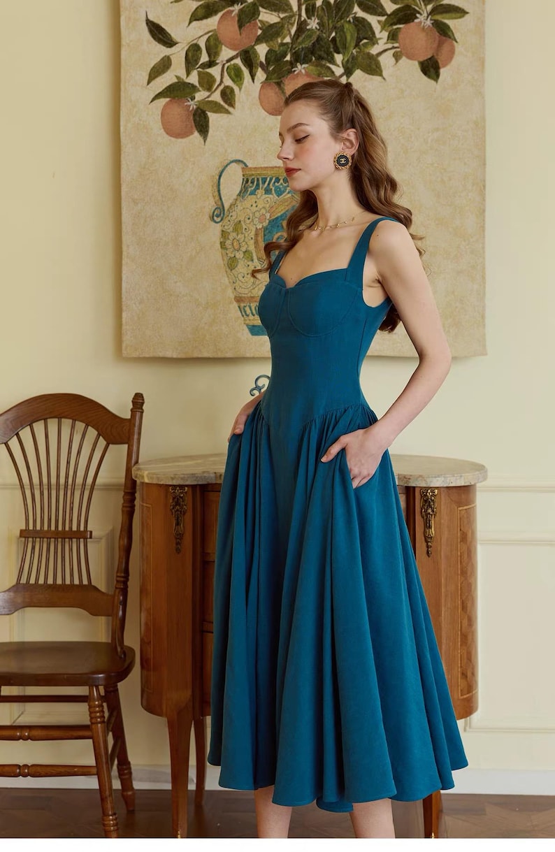 May include: A teal-colored dress with a sweetheart neckline and wide straps. The dress has a fitted bodice and a flared skirt with pockets. The model is standing in front of a painting of a vase and fruit.