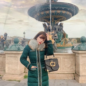 May include: A long, dark green puffer coat with a faux fur collar and gold and blue trim. The coat is worn with a black handbag with a gold clasp. The background features a large fountain.