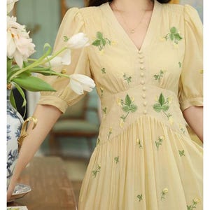Puede incluir: Vestido amarillo claro con mangas cortas abullonadas y escote en V. El vestido presenta delicados bordados florales verdes y amarillos en el corpi&ntilde;o y la falda. Tiene una fila de peque&ntilde;os botones en la parte delantera.
