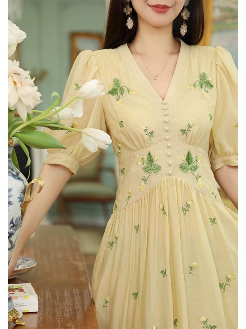Puede incluir: Vestido amarillo claro con mangas cortas abullonadas y escote en V. El vestido presenta delicados bordados florales verdes y amarillos en el corpi&ntilde;o y la falda. Tiene una fila de peque&ntilde;os botones en la parte delantera.