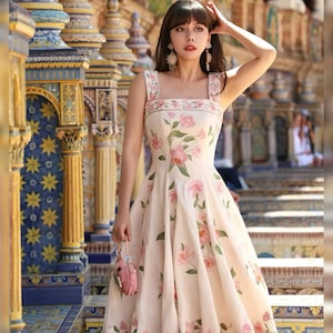 May include: A cream-coloured dress with a floral pattern of pink flowers and green leaves. The dress has a square neckline and a flared skirt. A small pink purse is held in the hand.