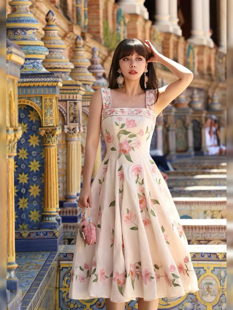 May include: A cream-coloured dress with a floral pattern of pink flowers and green leaves. The dress has a square neckline and a flared skirt. A small pink purse is held in the hand.