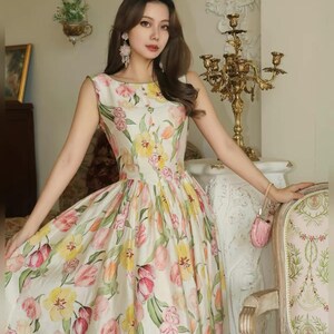 May include: A sleeveless, floor-length dress featuring a floral tulip design in pink, yellow, and green on a white base. The dress has a round neckline and a fitted bodice that flows into a full skirt. The dress is worn by a person.