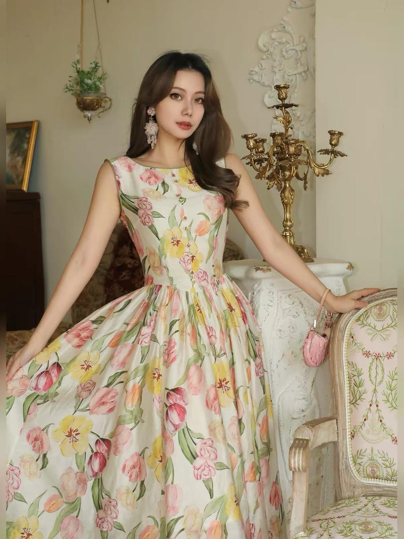 May include: A sleeveless, floor-length dress featuring a floral tulip design in pink, yellow, and green on a white base. The dress has a round neckline and a fitted bodice that flows into a full skirt. The dress is worn by a person.