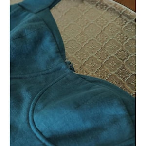 May include: Close-up of a teal-colored linen top with a scoop neckline and a structured bodice. The fabric has a subtle texture, and the garment appears to be well-tailored. The background features a patterned glass surface.