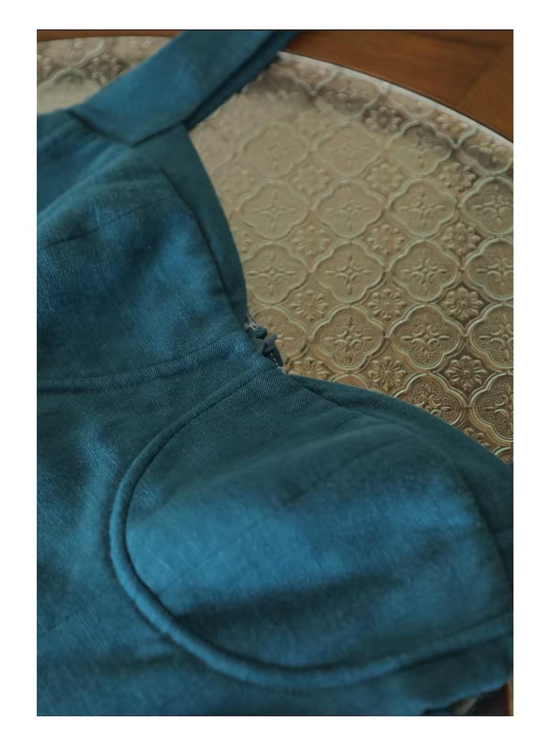 May include: Close-up of a teal-colored linen top with a scoop neckline and a structured bodice. The fabric has a subtle texture, and the garment appears to be well-tailored. The background features a patterned glass surface.