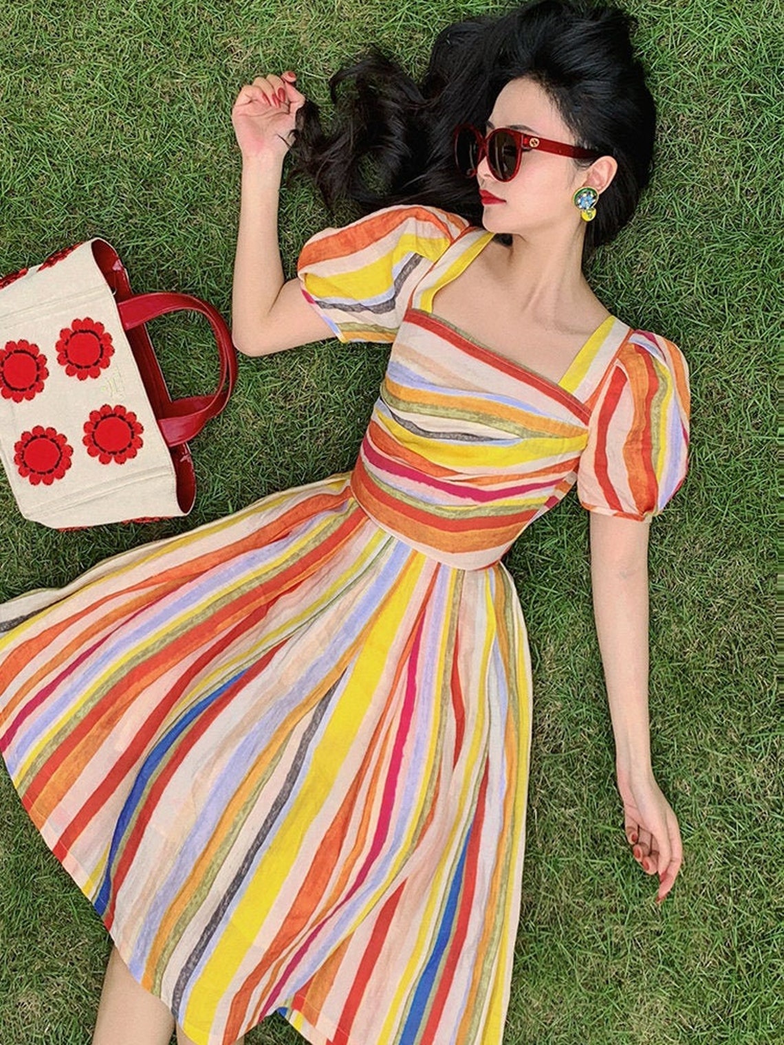 May include: A woman wearing a colorful striped dress with a red handbag. The dress has a square neckline and short puff sleeves. The woman is lying on her back in a grassy field.