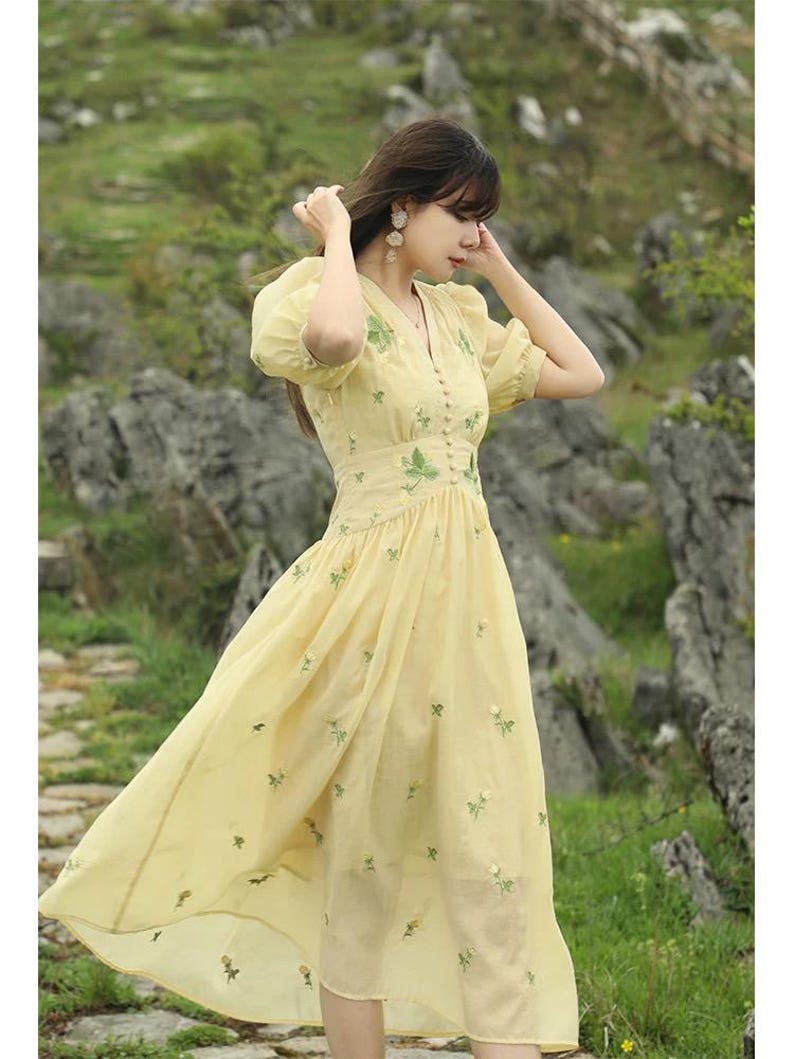 Puede incluir: Un vestido amarillo claro con mangas cortas abullonadas y cuello en V. El vestido presenta delicados bordados florales verdes y una falda fluida. El vestido est&aacute; hecho de una tela ligera. El vestido es de largo midi.
