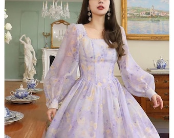 以下が含まれることがあります： 薄い紫色の花柄のロングスリーブドレス。スクエアネックラインとフィットしたボディス。スカートはボリュームがあり、流れるようなシルエットです。