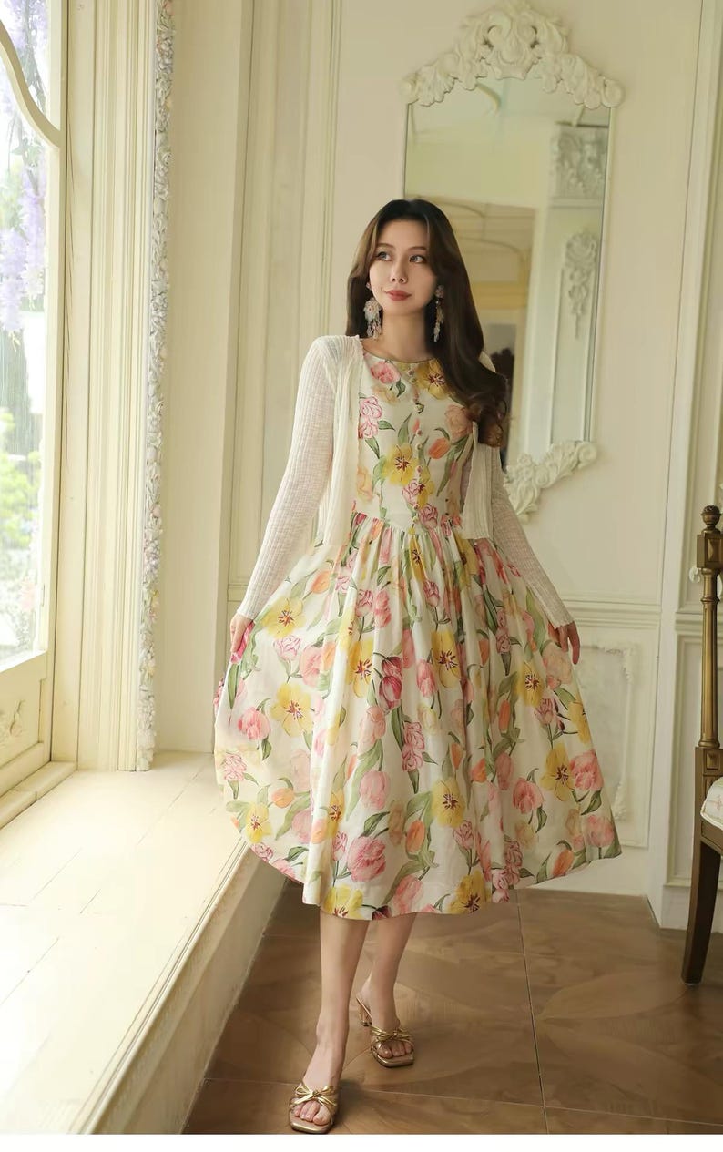 May include: A floral print dress with a white cardigan. The dress features a pattern of pink, yellow, and orange flowers on a cream background. The cardigan is a light, open-front style. The person is wearing gold sandals.