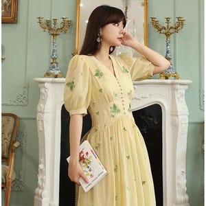 Puede incluir: Un vestido amarillo claro con mangas cortas abullonadas y cuello en V. El vestido presenta detalles de hojas bordadas en verde y peque&ntilde;os botones en la parte delantera. La persona sostiene un libro titulado "Flower Colour Theory".