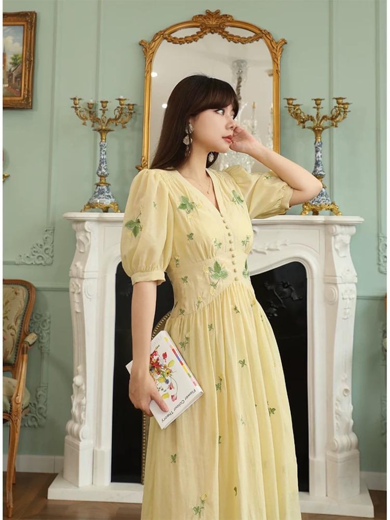 Puede incluir: Un vestido amarillo claro con mangas cortas abullonadas y cuello en V. El vestido presenta detalles de hojas bordadas en verde y peque&ntilde;os botones en la parte delantera. La persona sostiene un libro titulado "Flower Colour Theory".