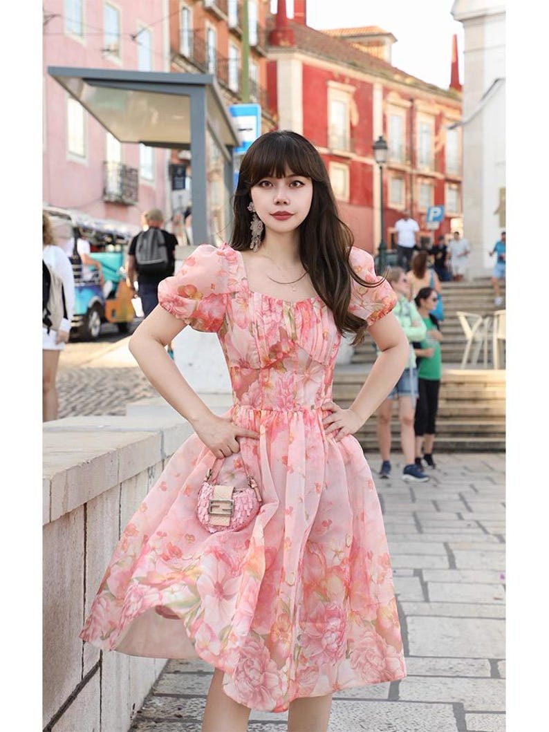 May include: A pink floral print dress with short puff sleeves and a square neckline. The dress has a fitted bodice and a flowing skirt. A small pink purse is held in the hand. The background shows a European street scene.