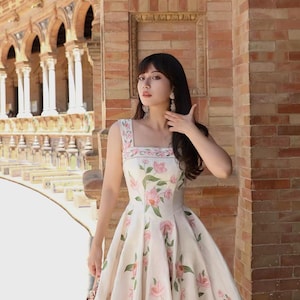 May include: A floral print dress with a square neckline. The dress is white with pink flowers and green leaves. The dress is knee-length and sleeveless. The image is taken outdoors in front of a brick building.