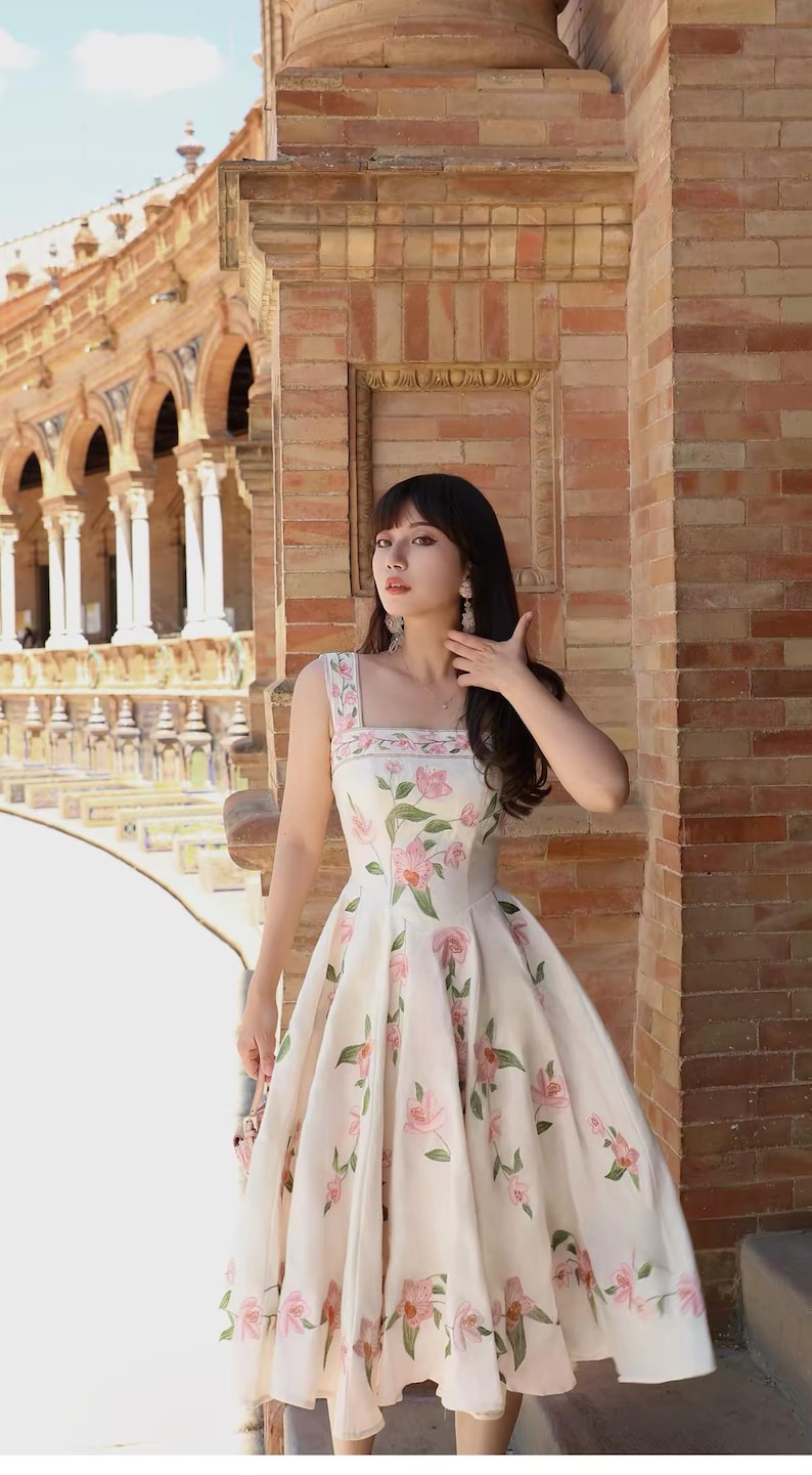 May include: A floral print dress with a square neckline. The dress is white with pink flowers and green leaves. The dress is knee-length and sleeveless. The image is taken outdoors in front of a brick building.