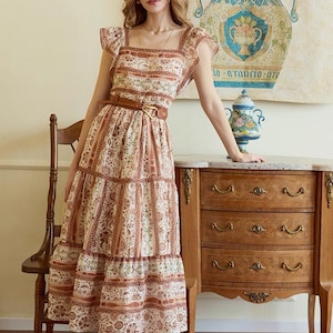 May include: A tiered, floral-print dress with a square neckline and flutter sleeves. The dress is cinched at the waist with a brown belt. The model is wearing gold sandals. A wooden chair and dresser are in the background.