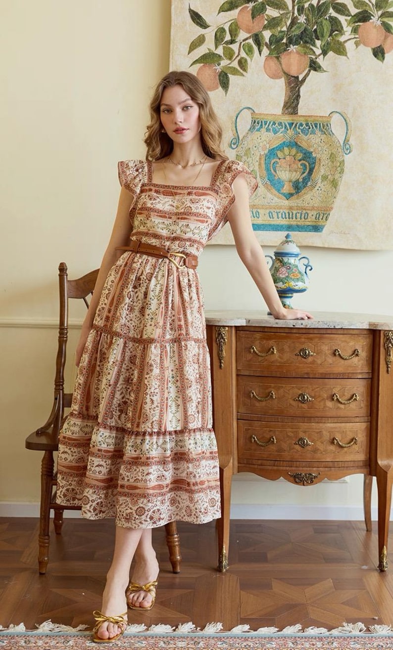 May include: A tiered, floral-print dress with a square neckline and flutter sleeves. The dress is cinched at the waist with a brown belt. The model is wearing gold sandals. A wooden chair and dresser are in the background.