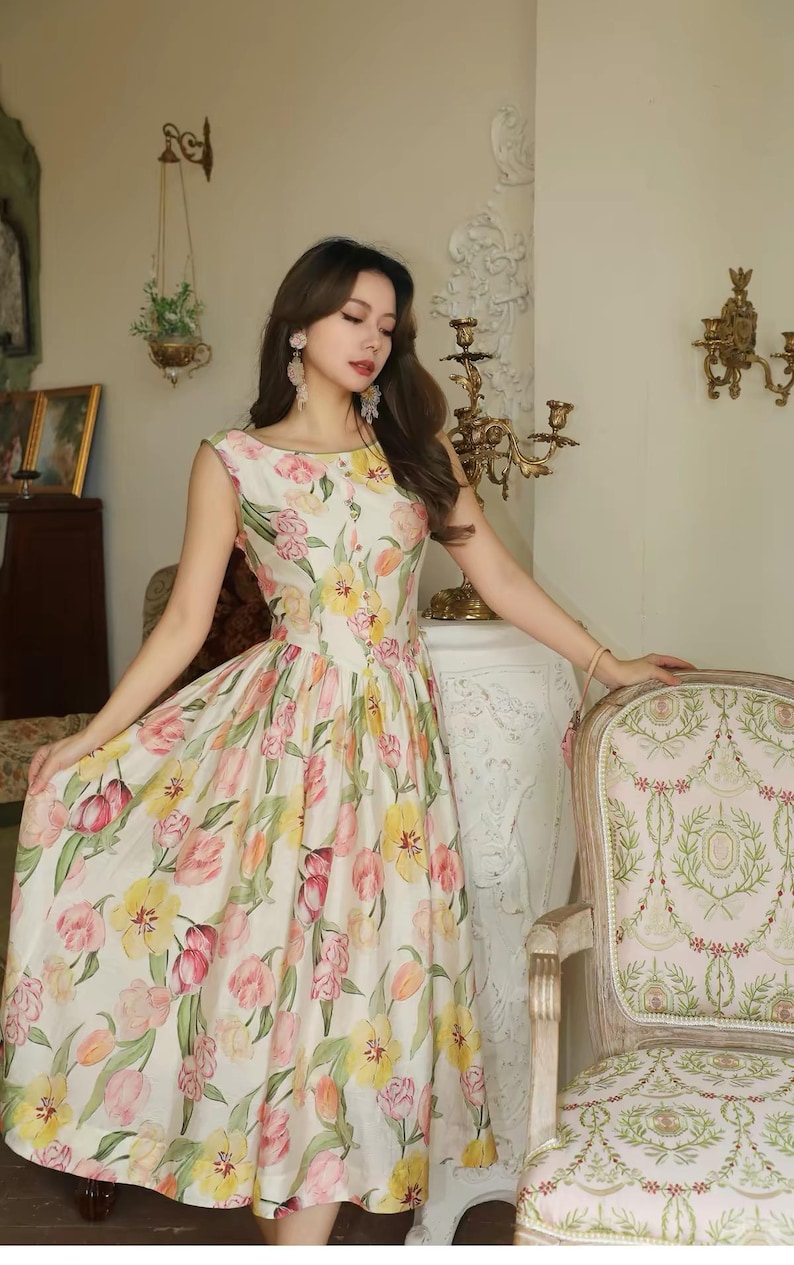 May include: A sleeveless dress with a floral print, featuring a scoop neckline and a full skirt. The dress displays pink, yellow, and orange tulips on a cream background. A vintage-style chair is also visible.