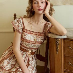 May include: A woman wearing a cream-colored dress with a floral and paisley pattern. The dress has short sleeves, a square neckline, and a brown belt. She is wearing gold jewelry and sitting near a wooden cabinet.