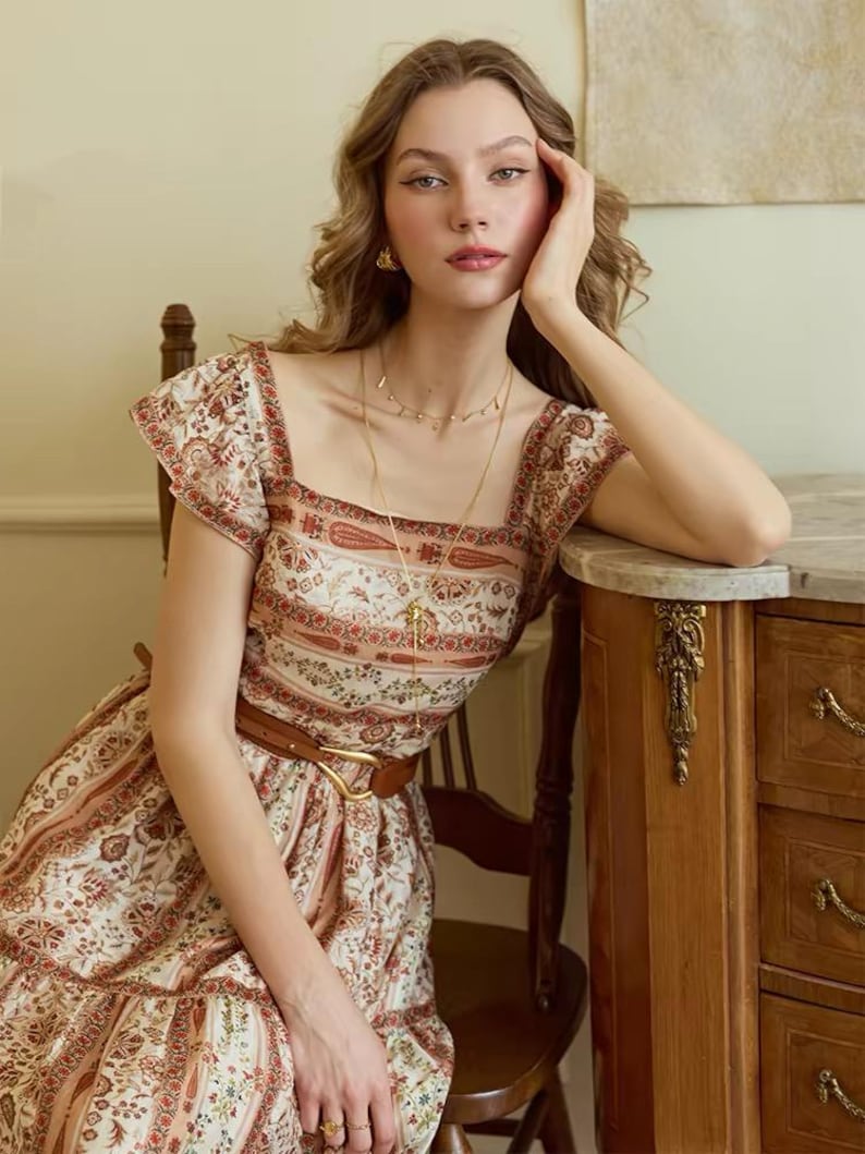 May include: A woman wearing a cream-colored dress with a floral and paisley pattern. The dress has short sleeves, a square neckline, and a brown belt. She is wearing gold jewelry and sitting near a wooden cabinet.