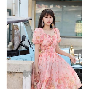 May include: A pink floral print dress with short, puffed sleeves and a square neckline. The dress features a full skirt and is paired with a small, pink handbag. The background includes a vintage blue car and a building.