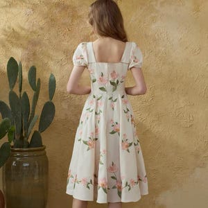 May include: A cream dress featuring a square neckline and short, puffed sleeves. The dress is decorated with pink floral patterns and green leaves. The model is standing in front of a textured, yellow-toned wall.