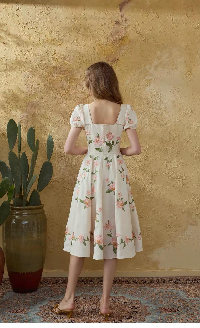 May include: A cream dress featuring a square neckline and short, puffed sleeves. The dress is decorated with pink floral patterns and green leaves. The model is standing in front of a textured, yellow-toned wall.