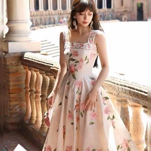 May include: A cream-coloured, sleeveless knee-length dress, decorated with pink floral embroidery. The dress has a square neckline and wide straps, with the floral pattern repeated on the bodice and skirt. The dress is made of a lightweight fabric, suitable for warmer weather.