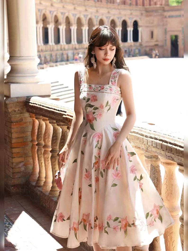 May include: A cream-coloured, sleeveless knee-length dress, decorated with pink floral embroidery. The dress has a square neckline and wide straps, with the floral pattern repeated on the bodice and skirt. The dress is made of a lightweight fabric, suitable for warmer weather.