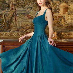 May include: A teal-colored dress with a fitted bodice and a flowing skirt. The dress features a sweetheart neckline and spaghetti straps. The model is standing in front of a tapestry with a landscape scene.