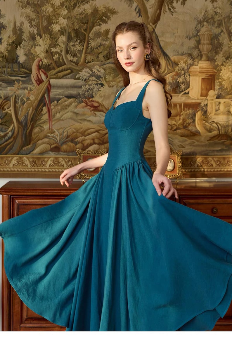 May include: A teal-colored dress with a fitted bodice and a flowing skirt. The dress features a sweetheart neckline and spaghetti straps. The model is standing in front of a tapestry with a landscape scene.