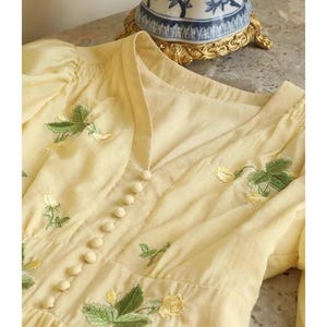 Puede incluir: Vestido amarillo p&aacute;lido con bordados florales. El vestido presenta un escote en V, peque&ntilde;os botones en la parte delantera y mangas abullonadas. Hojas verdes y flores amarillas bordadas en el corpi&ntilde;o y las mangas. El vestido es de tela ligera.