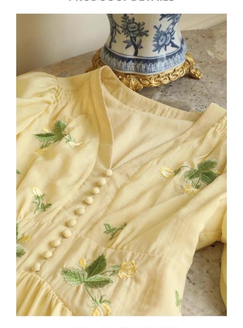 Puede incluir: Vestido amarillo p&aacute;lido con bordados florales. El vestido presenta un escote en V, peque&ntilde;os botones en la parte delantera y mangas abullonadas. Hojas verdes y flores amarillas bordadas en el corpi&ntilde;o y las mangas. El vestido es de tela ligera.