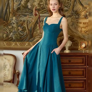 May include: A teal-colored dress with a sweetheart neckline and wide straps. The dress has a fitted bodice and a flowing skirt with pockets. The background features a tapestry and antique furniture, creating a vintage aesthetic.