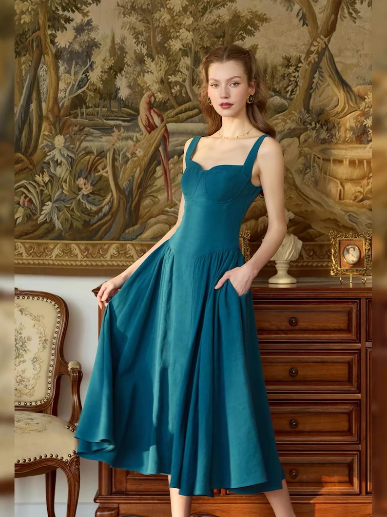 May include: A teal-colored dress with a sweetheart neckline and wide straps. The dress has a fitted bodice and a flowing skirt with pockets. The background features a tapestry and antique furniture, creating a vintage aesthetic.