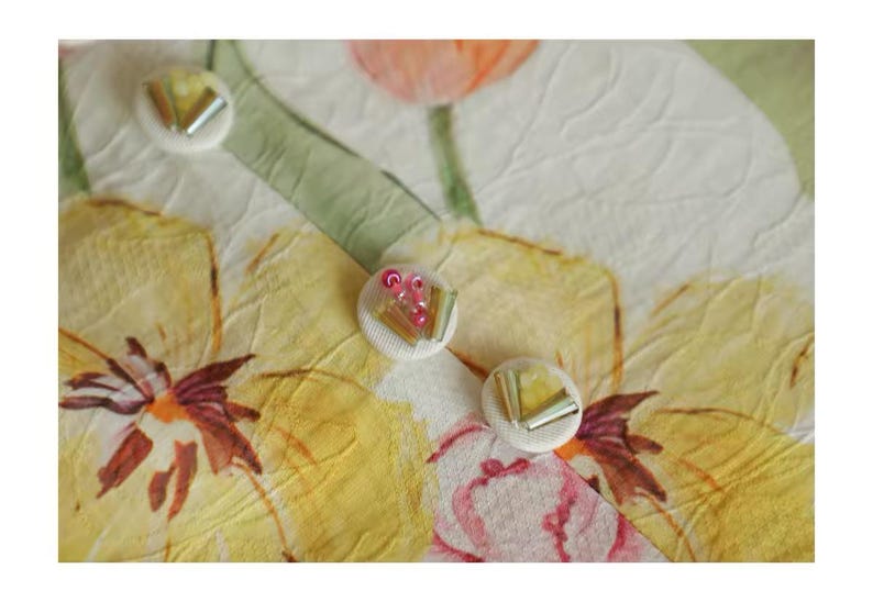 May include: Close-up of a floral pattern with three decorative buttons. The background features yellow, pink, and white flowers with green leaves. The buttons are white with silver and pink accents, adding a touch of elegance to the design.