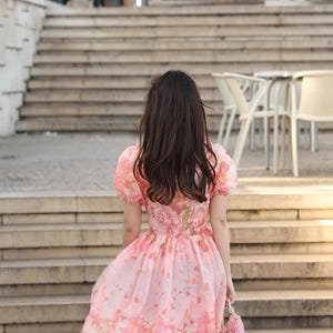 May include: A pink floral dress with short, puffed sleeves and a flowing skirt. The dress features a pattern of large pink flowers and is paired with a small, matching pink purse. The person is walking up a set of stone steps.