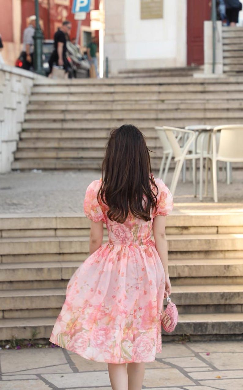 May include: A pink floral dress with short, puffed sleeves and a flowing skirt. The dress features a pattern of large pink flowers and is paired with a small, matching pink purse. The person is walking up a set of stone steps.