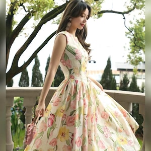 May include: A cream-coloured, sleeveless dress with a floral print in pink and yellow. The dress has a fitted bodice, a flared skirt, and a button-down front. A small pink purse is held.