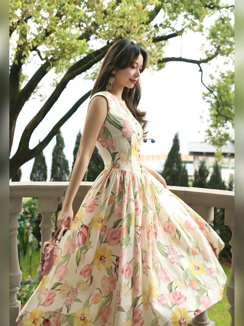 May include: A cream-coloured, sleeveless dress with a floral print in pink and yellow. The dress has a fitted bodice, a flared skirt, and a button-down front. A small pink purse is held.