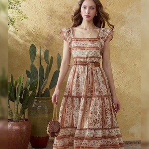 May include: A patterned, tiered dress in cream and brown tones. The dress features a square neckline, short sleeves, and a brown belt. A small, woven handbag is held in the hand. The background includes potted plants and a textured wall.