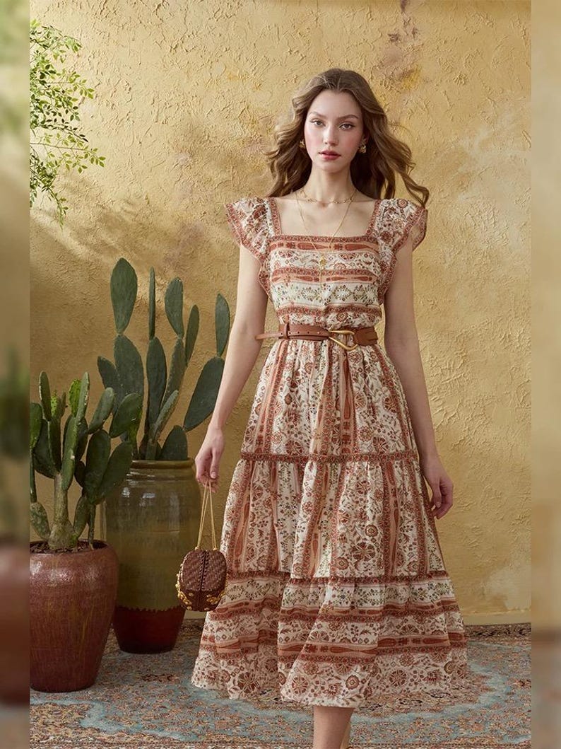 May include: A patterned, tiered dress in cream and brown tones. The dress features a square neckline, short sleeves, and a brown belt. A small, woven handbag is held in the hand. The background includes potted plants and a textured wall.
