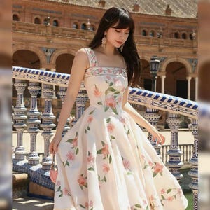 May include: A cream-coloured dress with a floral pattern of pink flowers and green leaves. The dress has a square neckline, wide straps, and a flared skirt. The dress is a midi length.