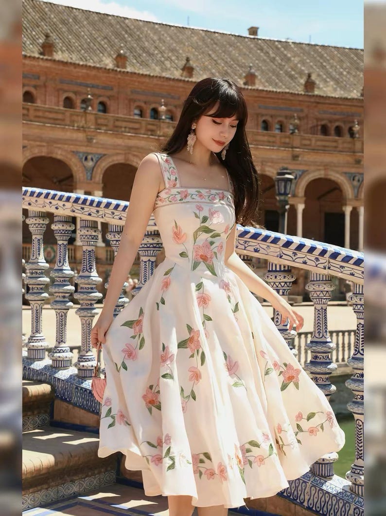 May include: A cream-coloured dress with a floral pattern of pink flowers and green leaves. The dress has a square neckline, wide straps, and a flared skirt. The dress is a midi length.