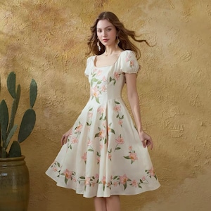 May include: A cream-coloured dress adorned with a floral design of pink flowers and green leaves. The dress features a square neckline, short puff sleeves, and a knee-length skirt. The model wears gold sandals.