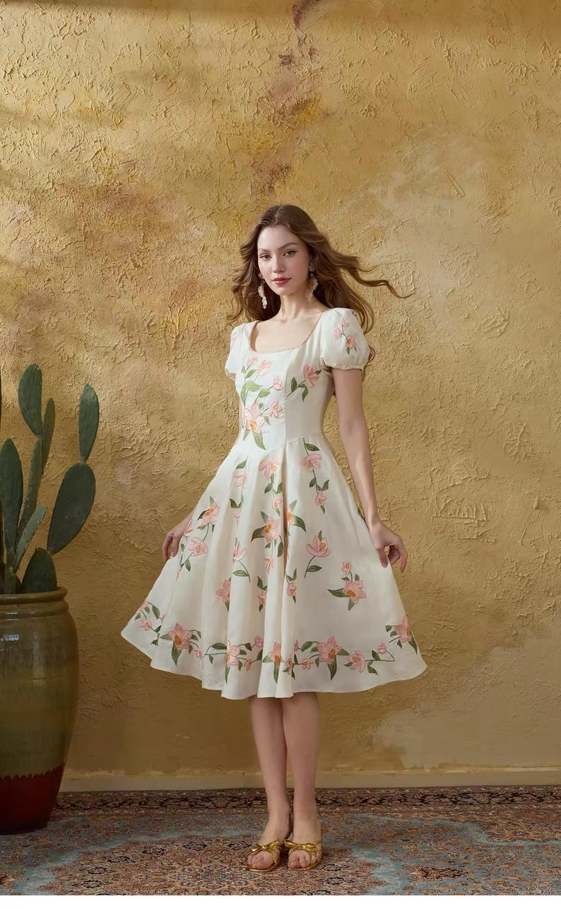May include: A cream-coloured dress adorned with a floral design of pink flowers and green leaves. The dress features a square neckline, short puff sleeves, and a knee-length skirt. The model wears gold sandals.
