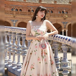 May include: A cream dress adorned with a floral design of pink flowers and green foliage. The dress features a square neckline and broad straps. A small pink purse is held. The backdrop showcases a building with arches and decorative columns.